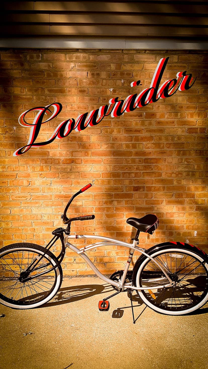 Silver lowrider bicycle with whitewall tires parked against a brick wall with stylized red lettering.