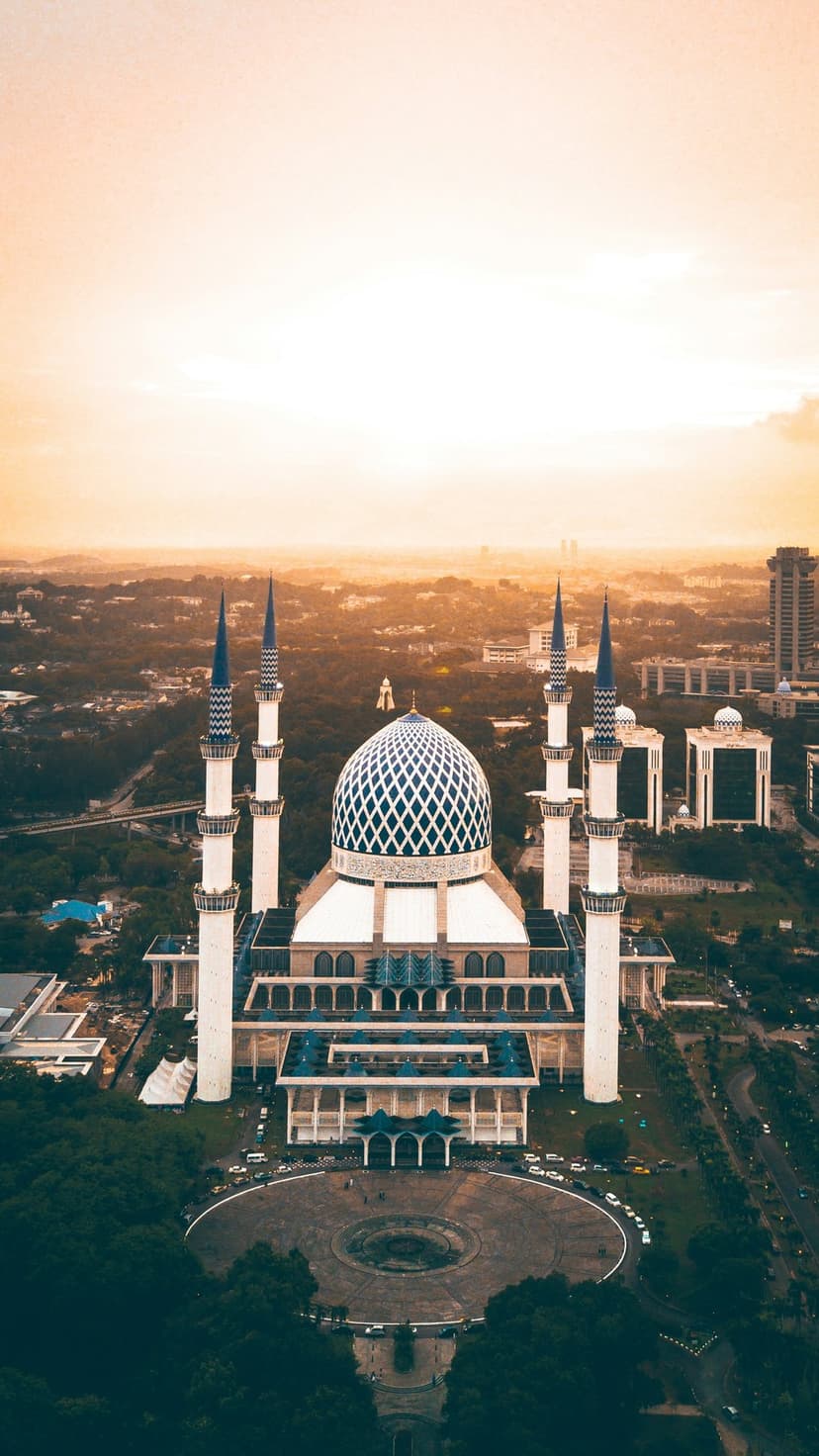 Sultan Salahuddin Abdul Aziz Mosque features a blue dome and minarets during a warm golden hour aerial sunset view.