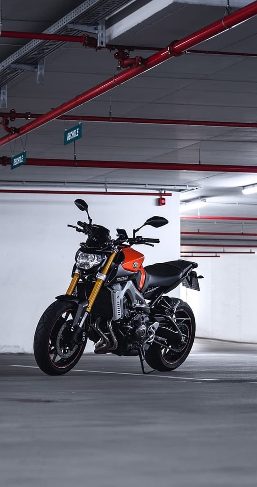 Orange and black Yamaha sportbike parked on concrete in a modern garage with overhead lights and red ceiling pipes.
