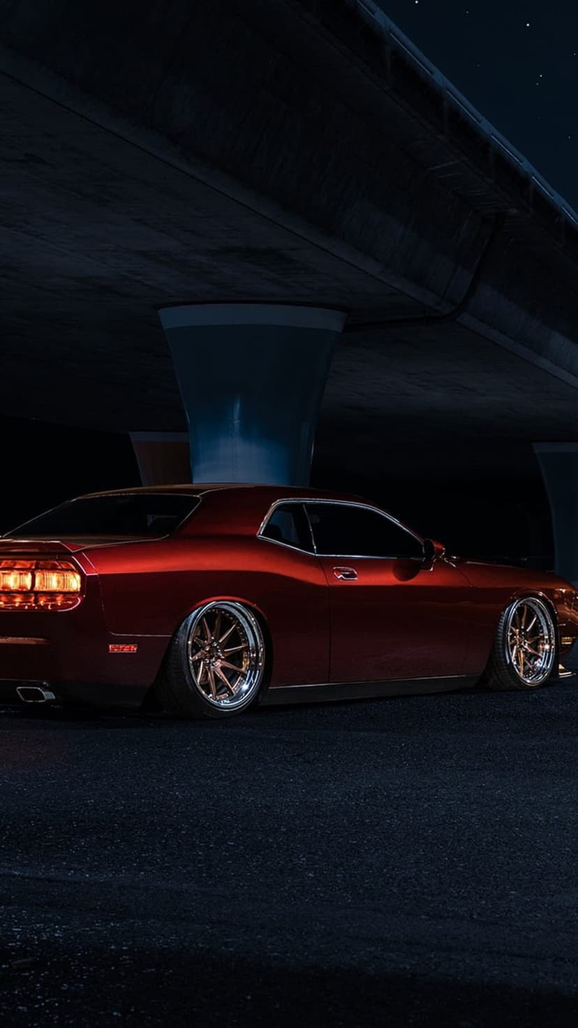 Red Dodge Challenger with bronze rims and a lowered suspension parked at night under a concrete overpass.