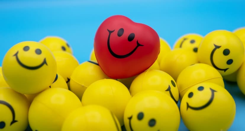 Red heart-shaped smiley ball resting on top of yellow round smiley face toys against a blue sky background.