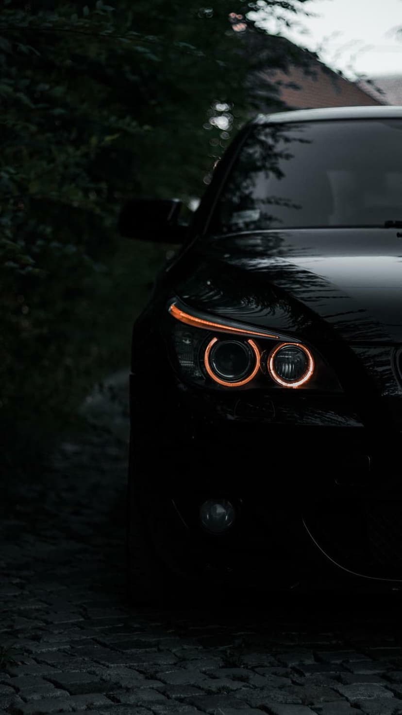 BMW headlights with orange circular LEDs glow on a dark cobblestone path framed by distant trees at night.