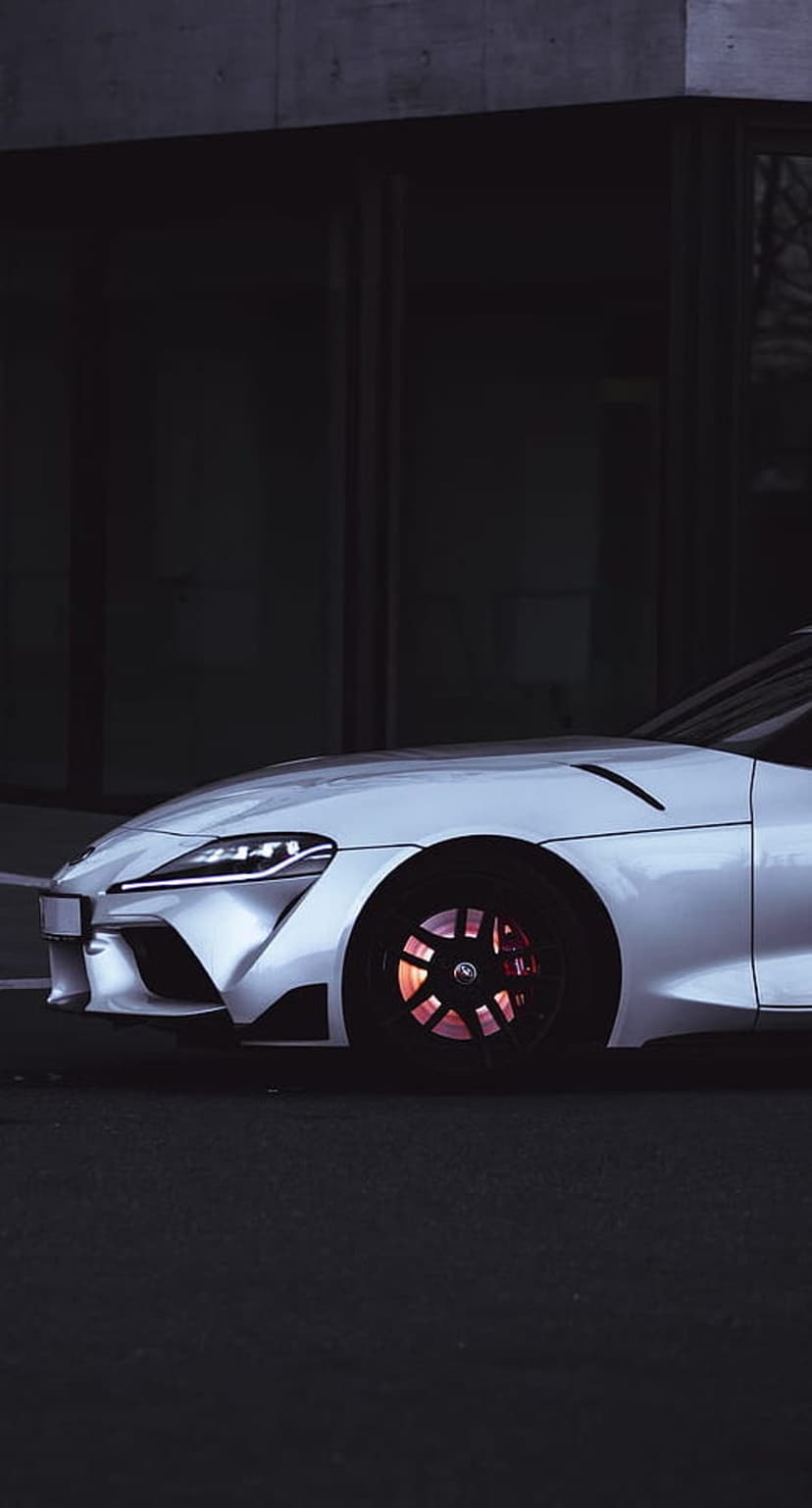 White sports car front wheel featuring red brake calipers against a dark building at a low angle perspective.
