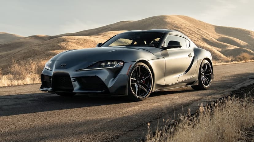 Silver Toyota Supra parked on a paved road with golden rolling hills in the background during a soft sunset.