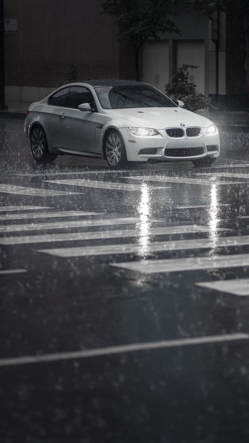 BMW M3 white coupe cruises over a rain-slicked crosswalk with glowing headlights and vibrant pavement reflections.