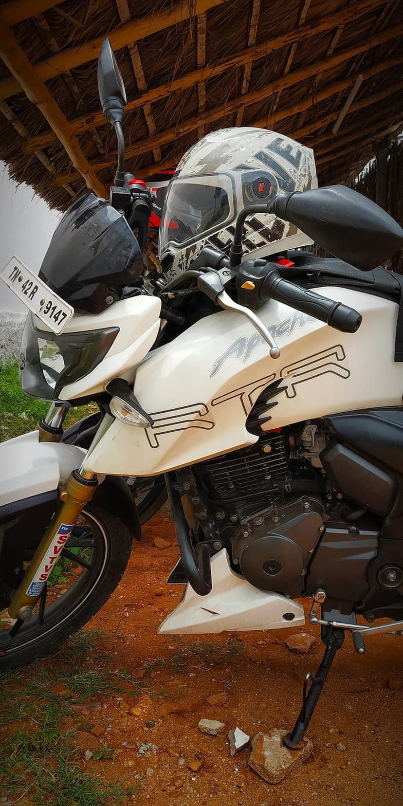 White Apache RTR motorcycle with a patterned helmet on the handlebars under a thatched roof on dirt ground.