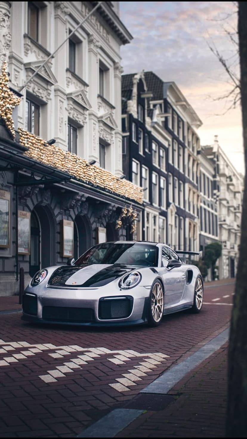 Silver Porsche GT2 RS with racing stripes on Amsterdam cobblestones near glowing historic canal houses.