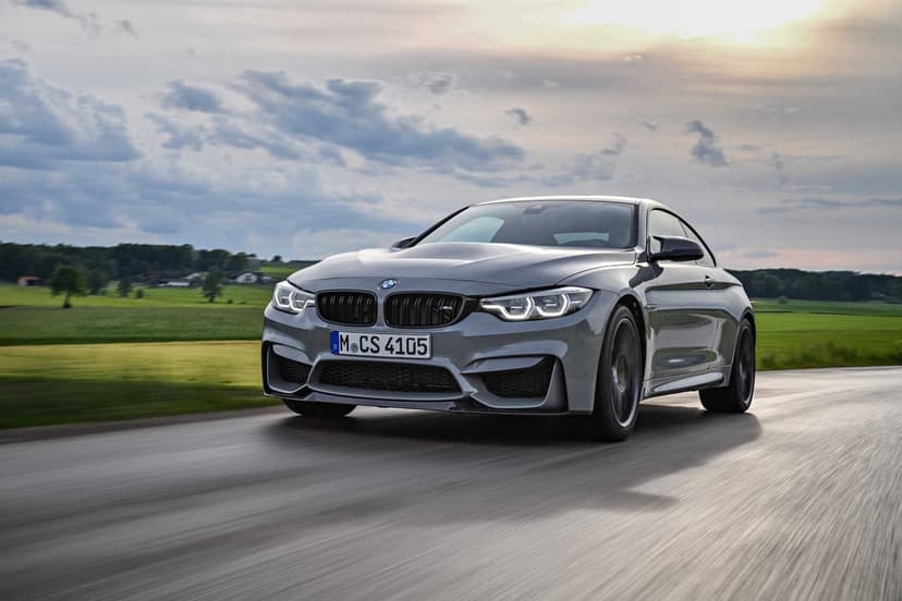 Grey BMW M4 CS driving on a blurred rural road with green fields and a cloudy sunset sky in the background.