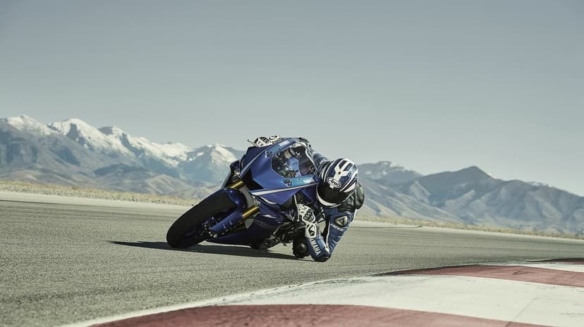 Blue Yamaha sportbike leans deep into a racetrack turn with snow-capped mountains under a clear sky.