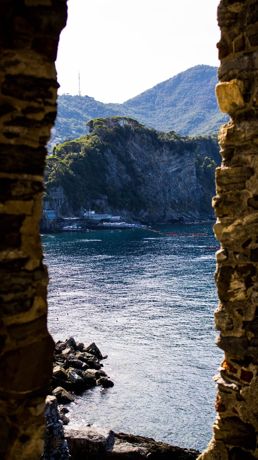 Weathered stone archway framing a view of green Italian hills and the blue Mediterranean sea under bright sunlight.