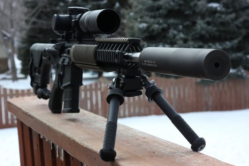 AR-15 rifle with a high-powered scope and suppressor rests on a wooden fence rail against a snowy winter backdrop.