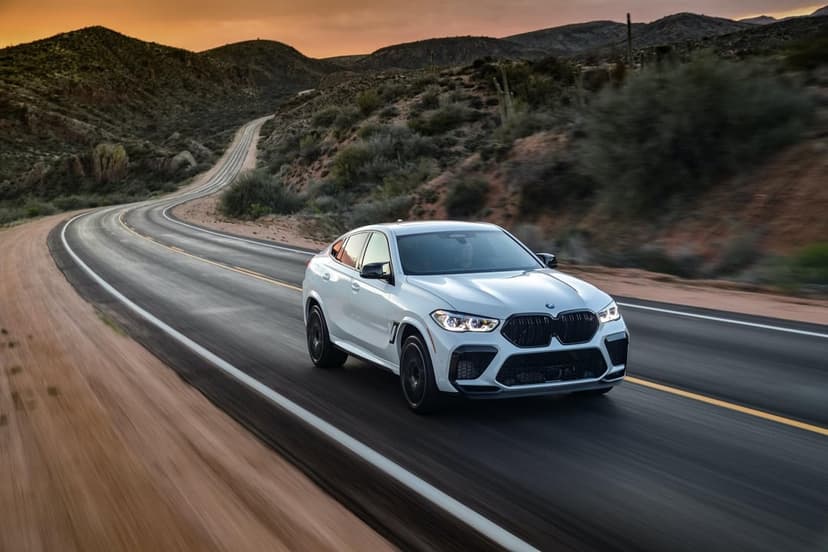 White BMW X6 M driving on a winding desert road during a golden sunset with mountains in the background.