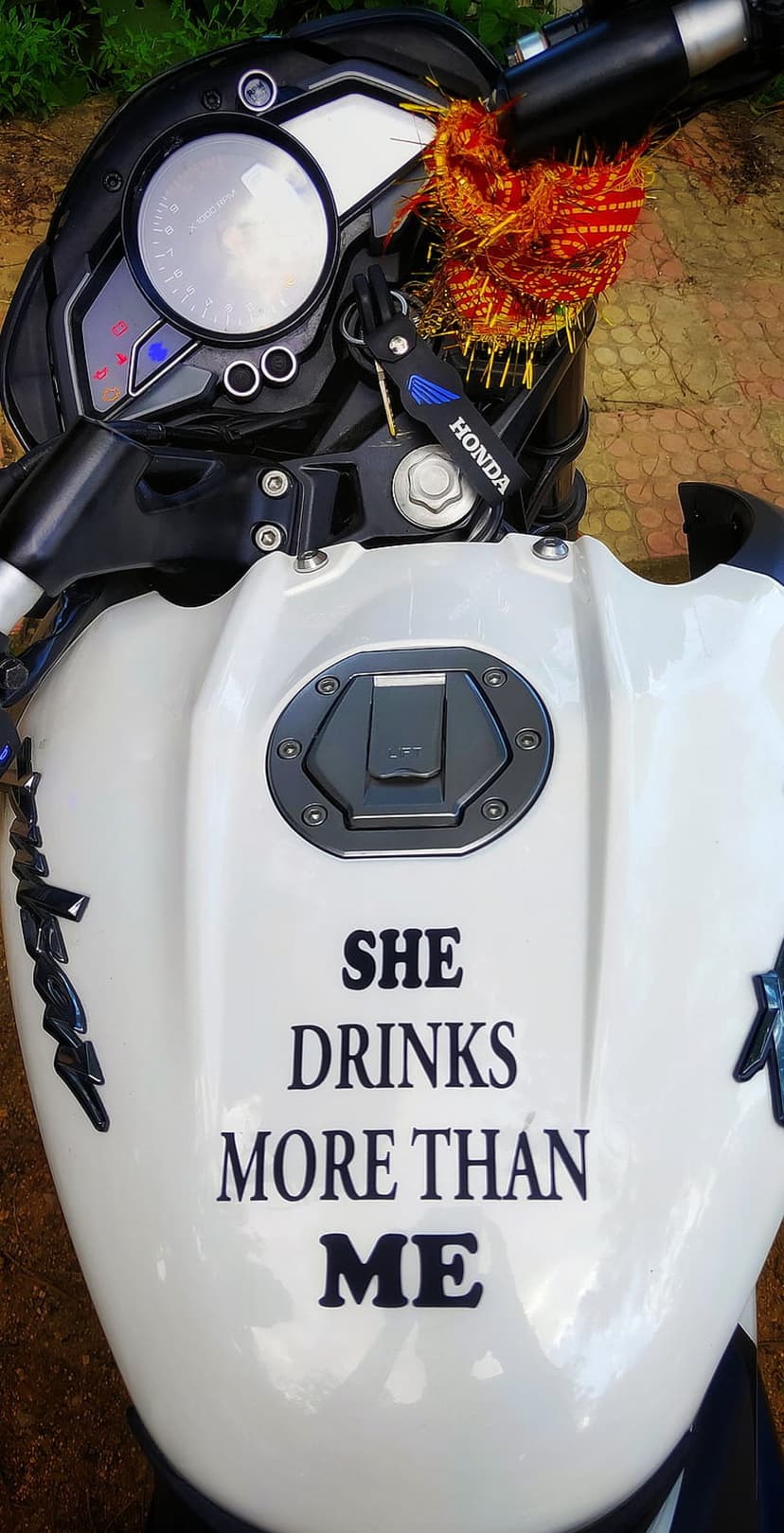 White Honda fuel tank with a black sticker reading She Drinks More Than Me and a red prayer thread on the key.