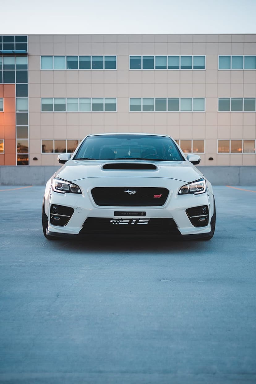 White Subaru WRX STI with a large hood scoop parked on concrete in front of a modern glass office building.