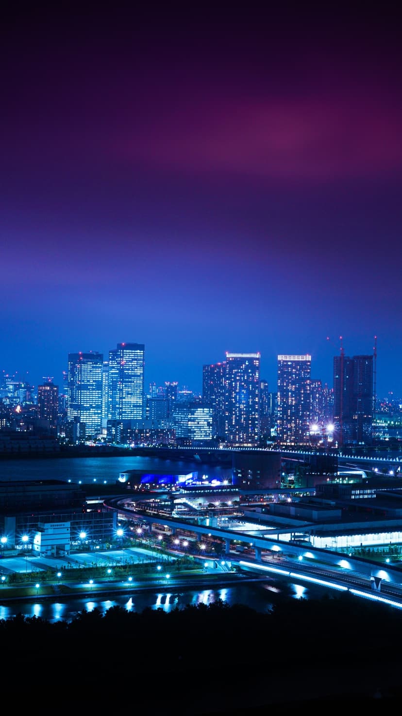 Illuminated skyscrapers tower over a dark harbor with vibrant purple and blue reflections on the water's surface.