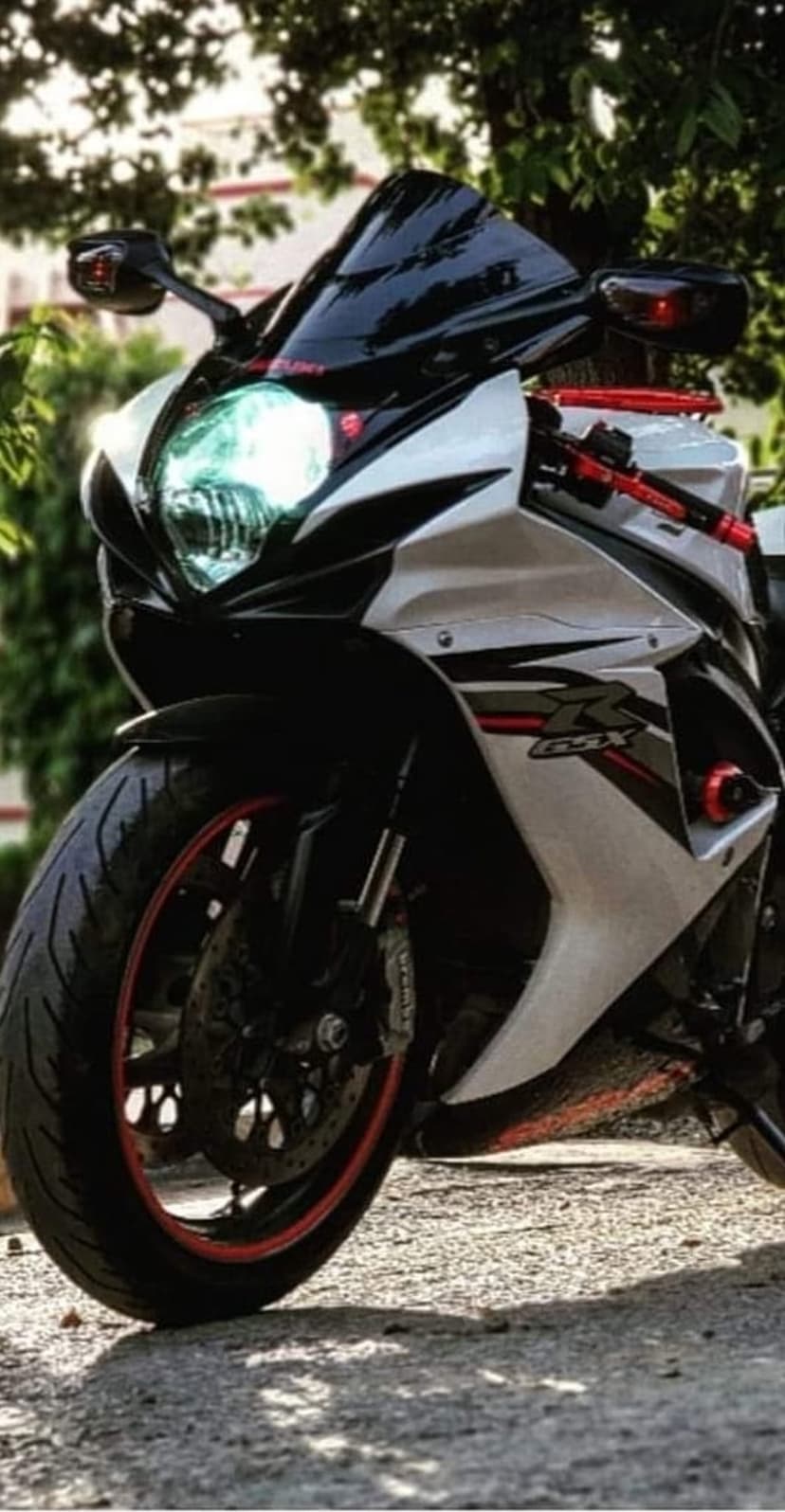 Suzuki GSX-R sportbike with white fairings and red rim accents parked on a textured path near green bushes.
