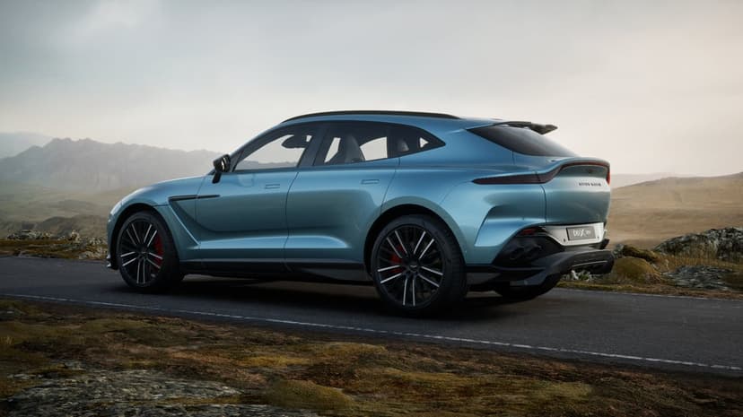 Teal Aston Martin DBX 707 SUV on a winding road with mountains and a cloudy sky in the background.