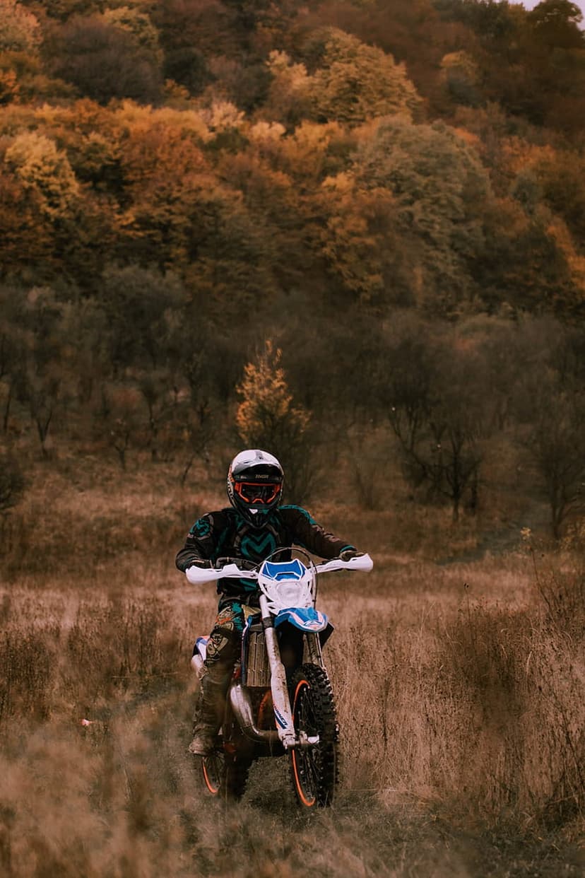 Motocross rider sits on a dirt bike in a dry grassy field framed by vibrant orange autumn forest foliage.