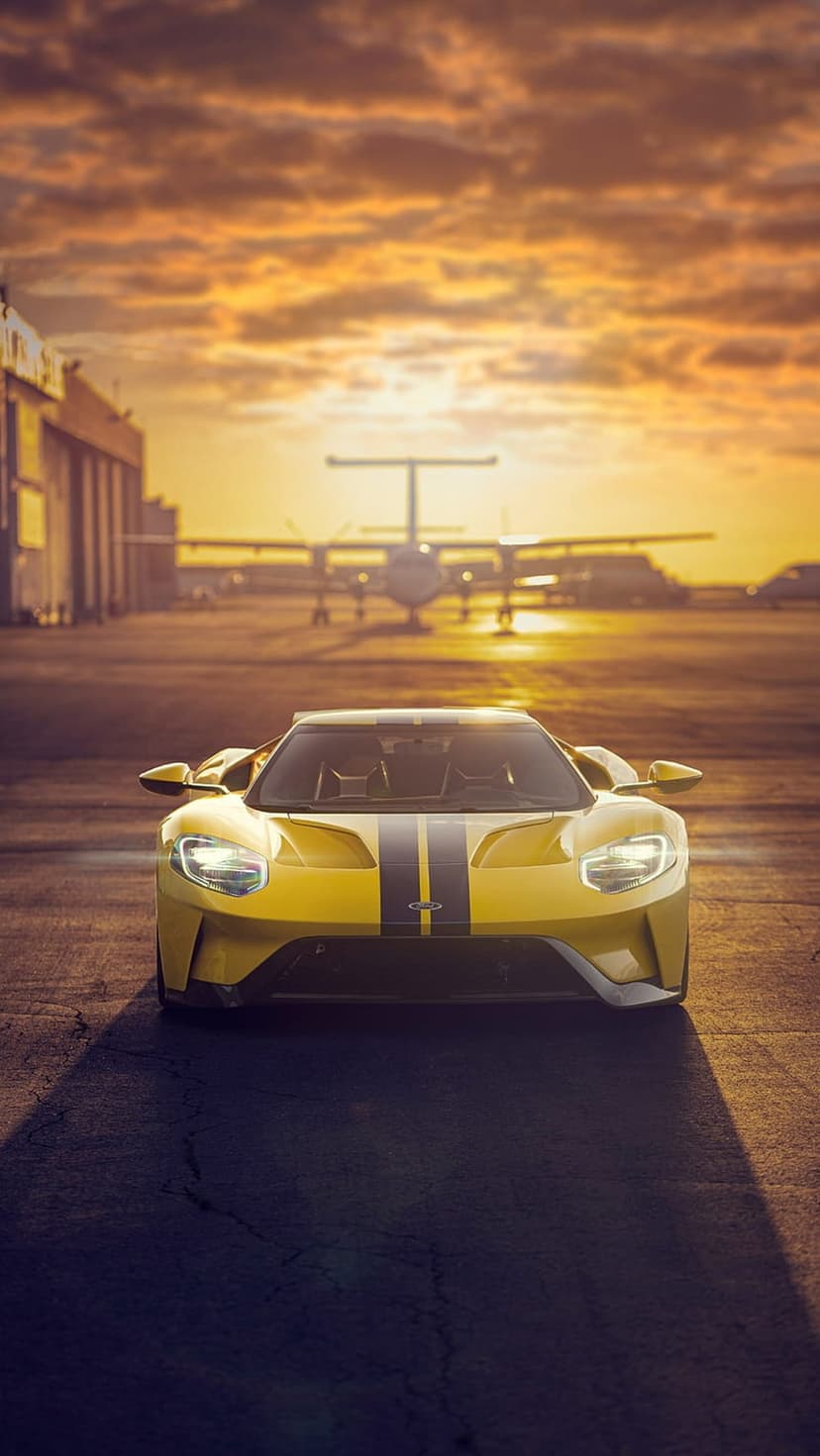 Yellow Ford GT supercar on an airport runway beneath a fiery orange sunset sky with a distant airplane.