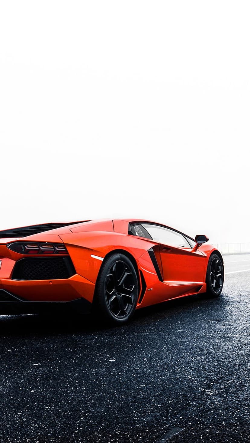 Orange Lamborghini Aventador parked on wet asphalt at a low angle showing its rear profile and black wheels.