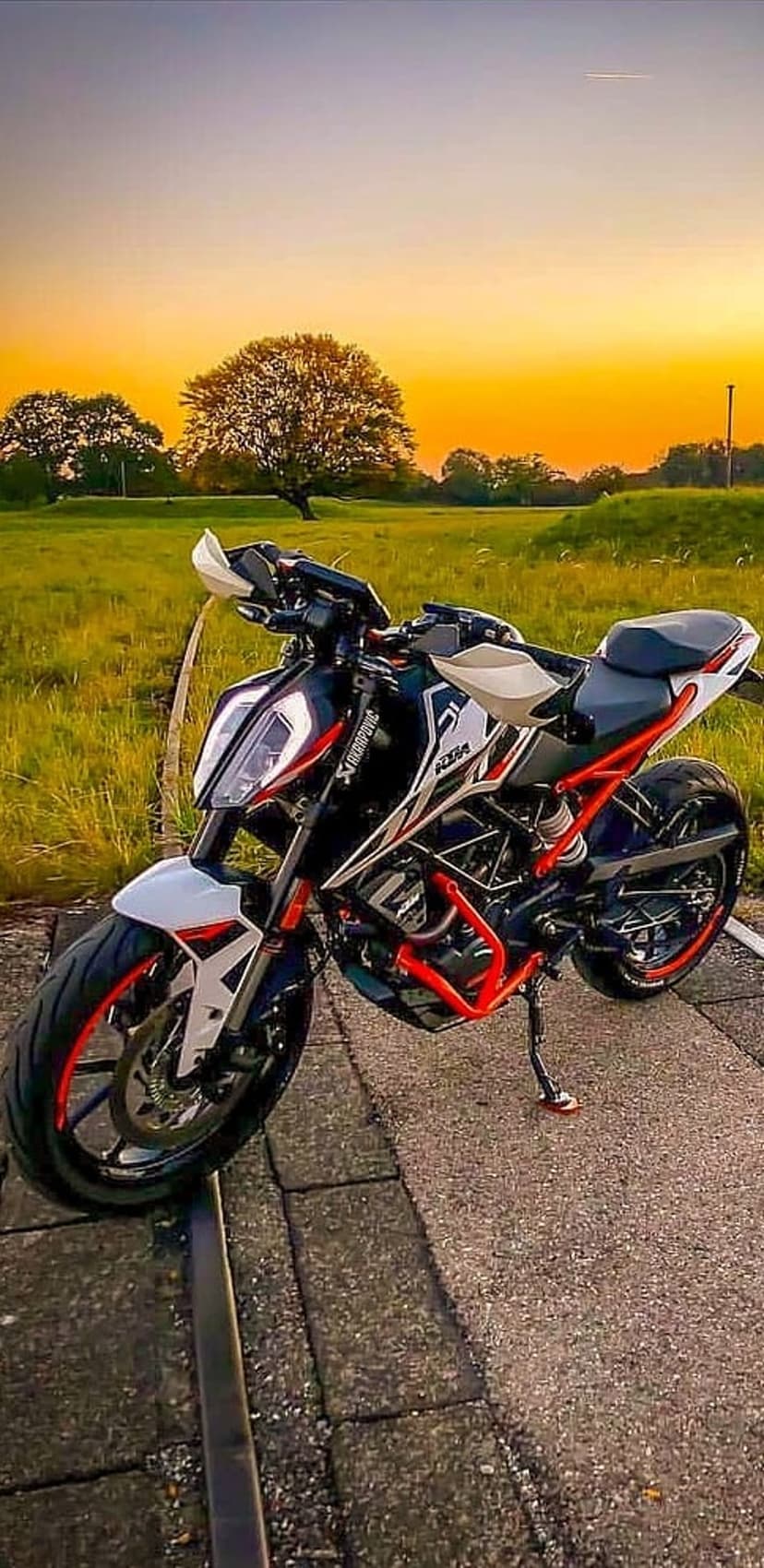 KTM motorcycle parked on an asphalt path against a blazing orange sunset and silhouetted trees in a field.