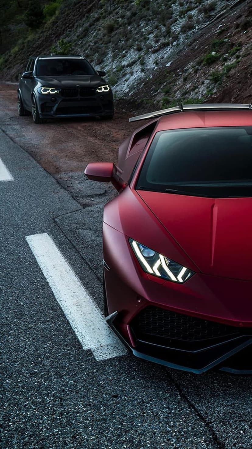 Matte red Lamborghini and black BMW SUV parked on a mountain road with headlights on against a rocky hillside.