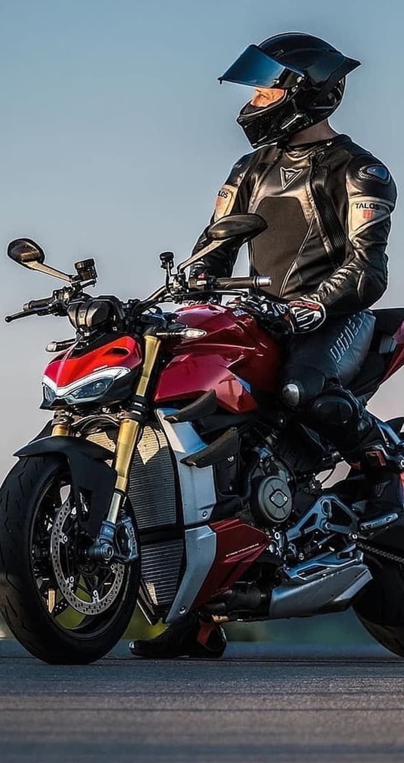 Motorcyclist in black leathers sits on a red Ducati bike featuring golden forks under a bright clear sky.