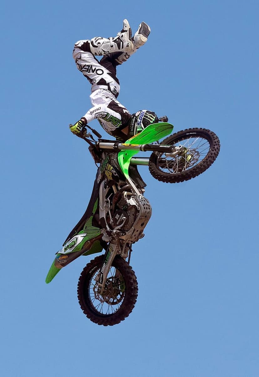 Motocross rider in full gear performs an inverted mid-air stunt on a green dirt bike against a clear blue sky.