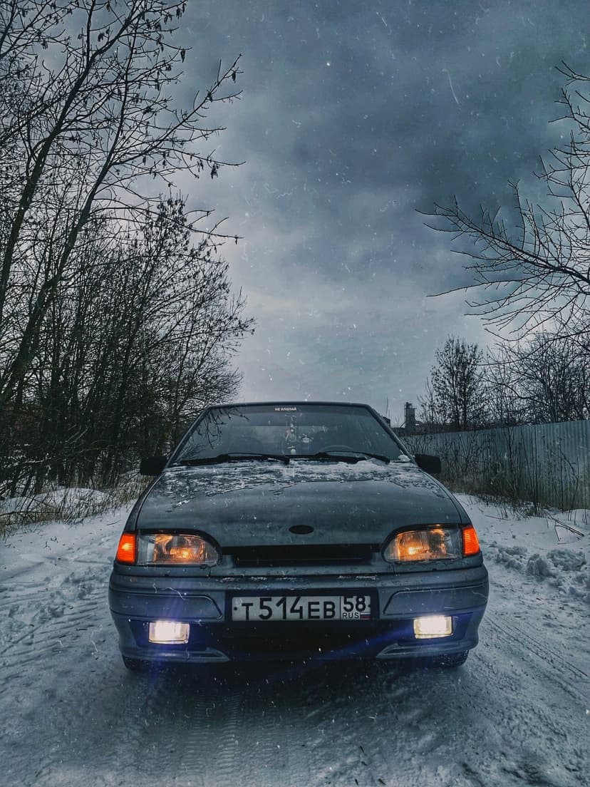 Dark grey car with glowing headlights on a snowy road surrounded by bare trees during a heavy winter blizzard.