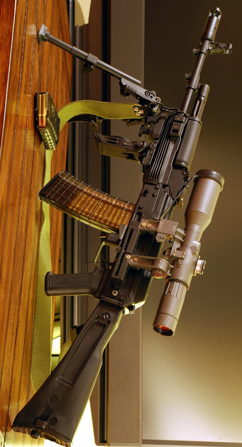 Black assault rifle with a mounted scope and folded bipod standing vertically against a natural wood background.