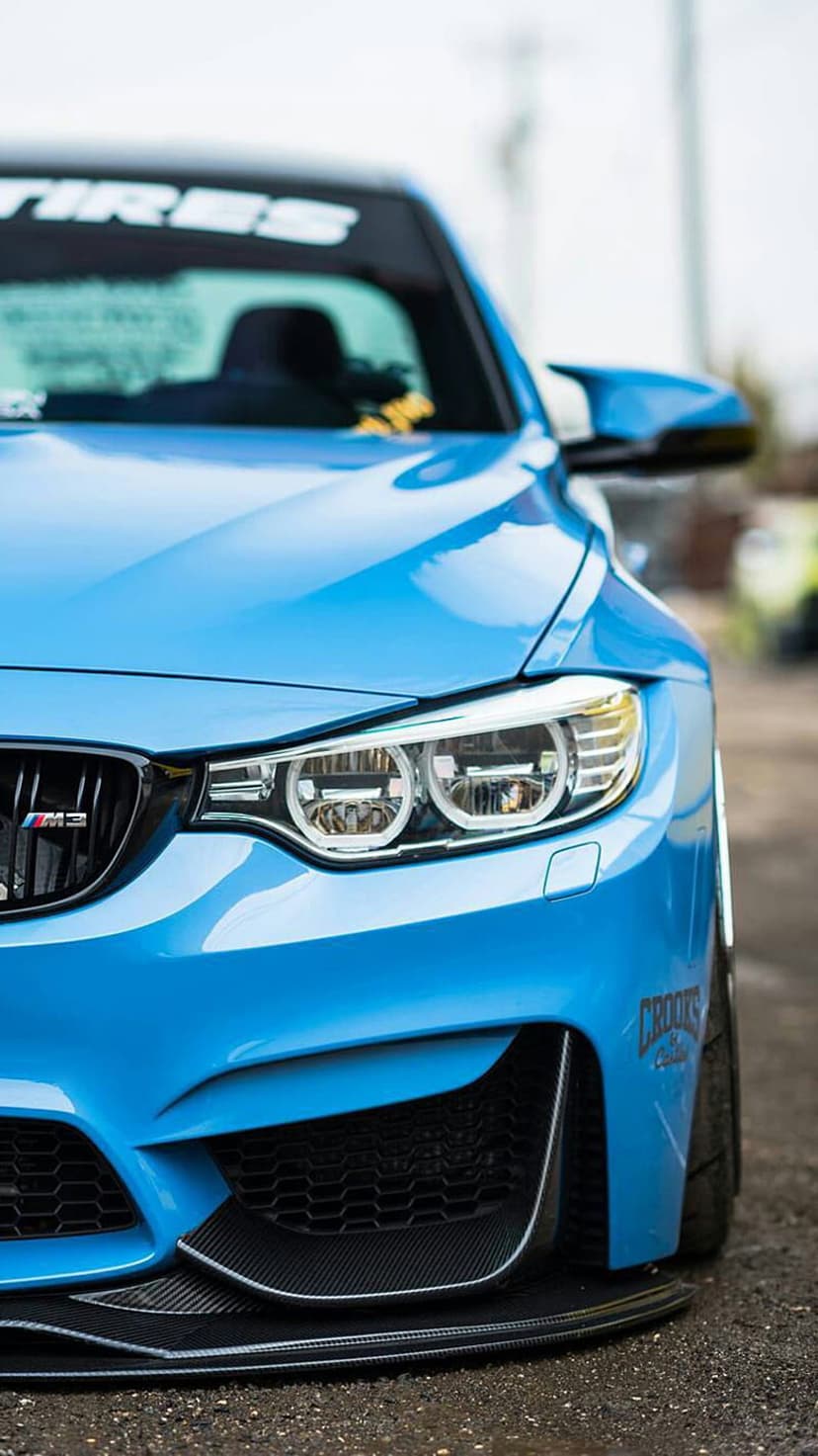 Blue BMW M4 Coupe featuring a carbon fiber front splitter and honeycomb grille on a sunlit asphalt road.