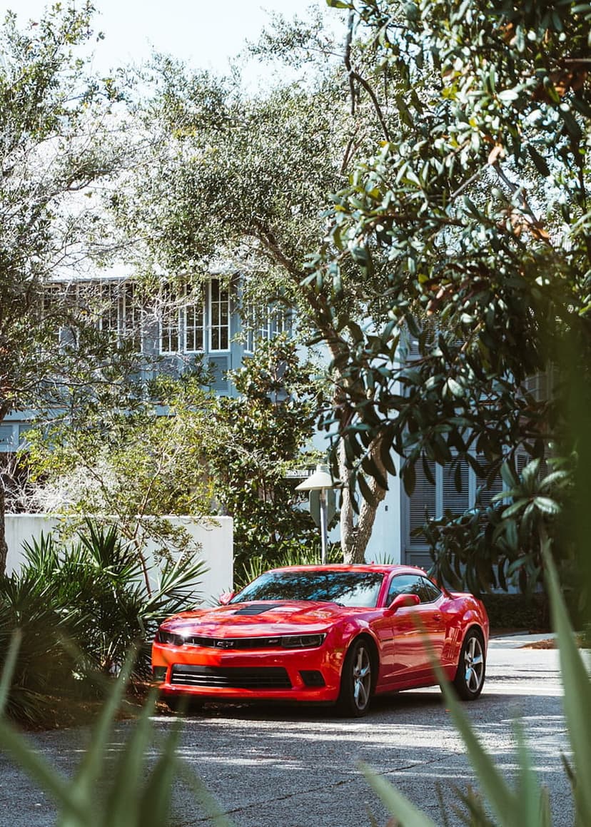Red Chevrolet Camaro SS parked on a stone driveway next to a modern gray house with large glass windows and trees.