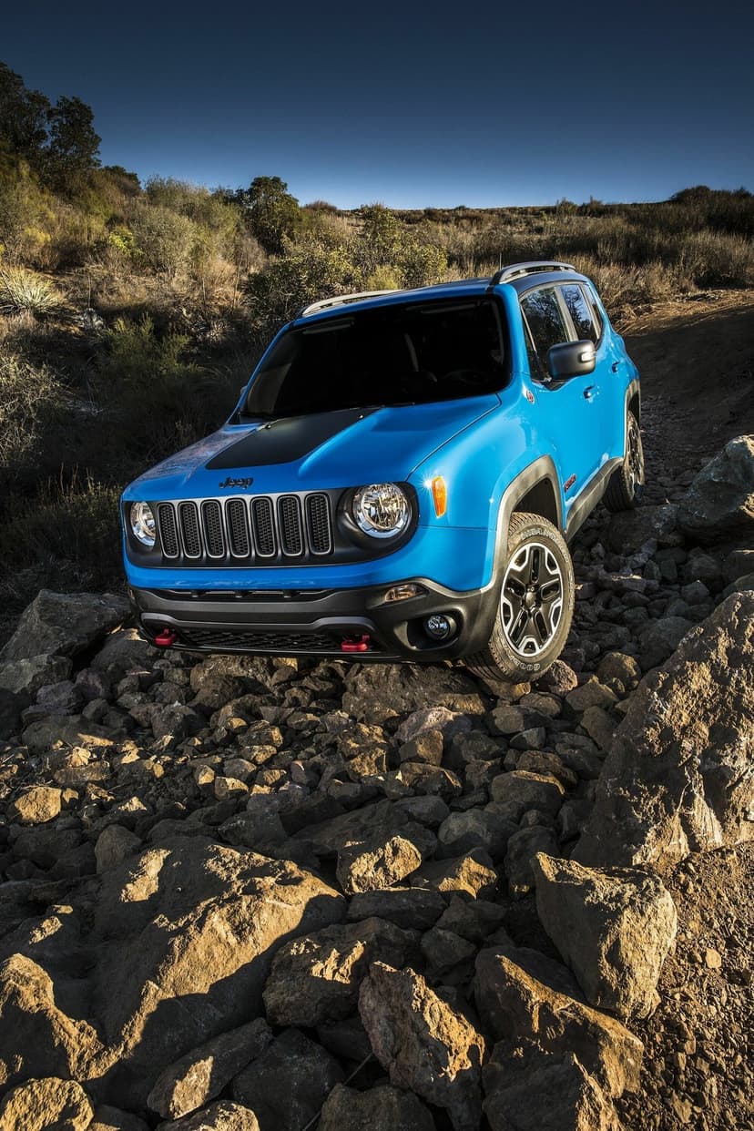 Vibrant blue Jeep Renegade Trailhawk climbs over large brown boulders on a rugged off-road mountain trail.