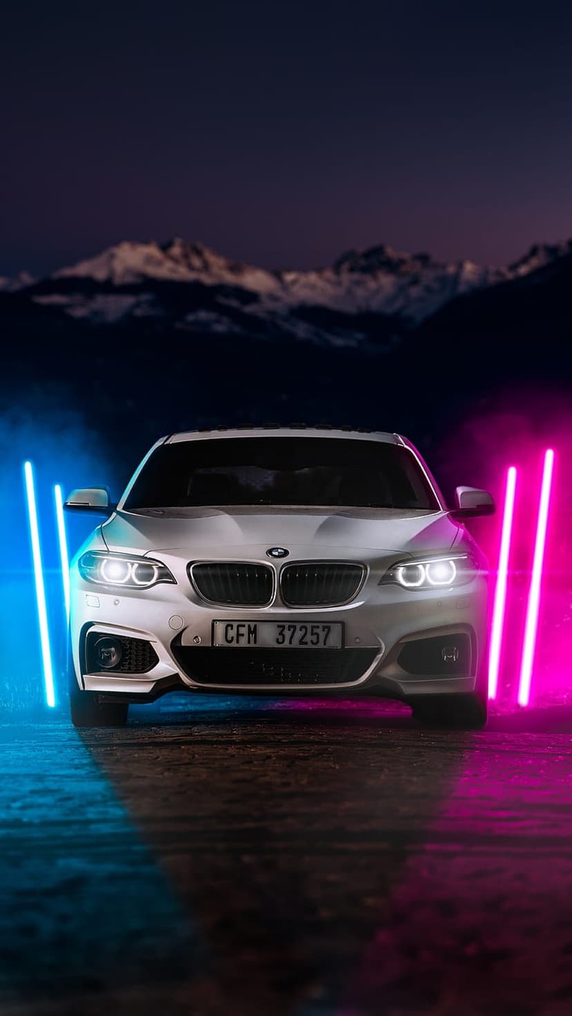 White BMW car illuminated by pink and blue neon lights on a snowy mountain road during the blue hour at dusk.