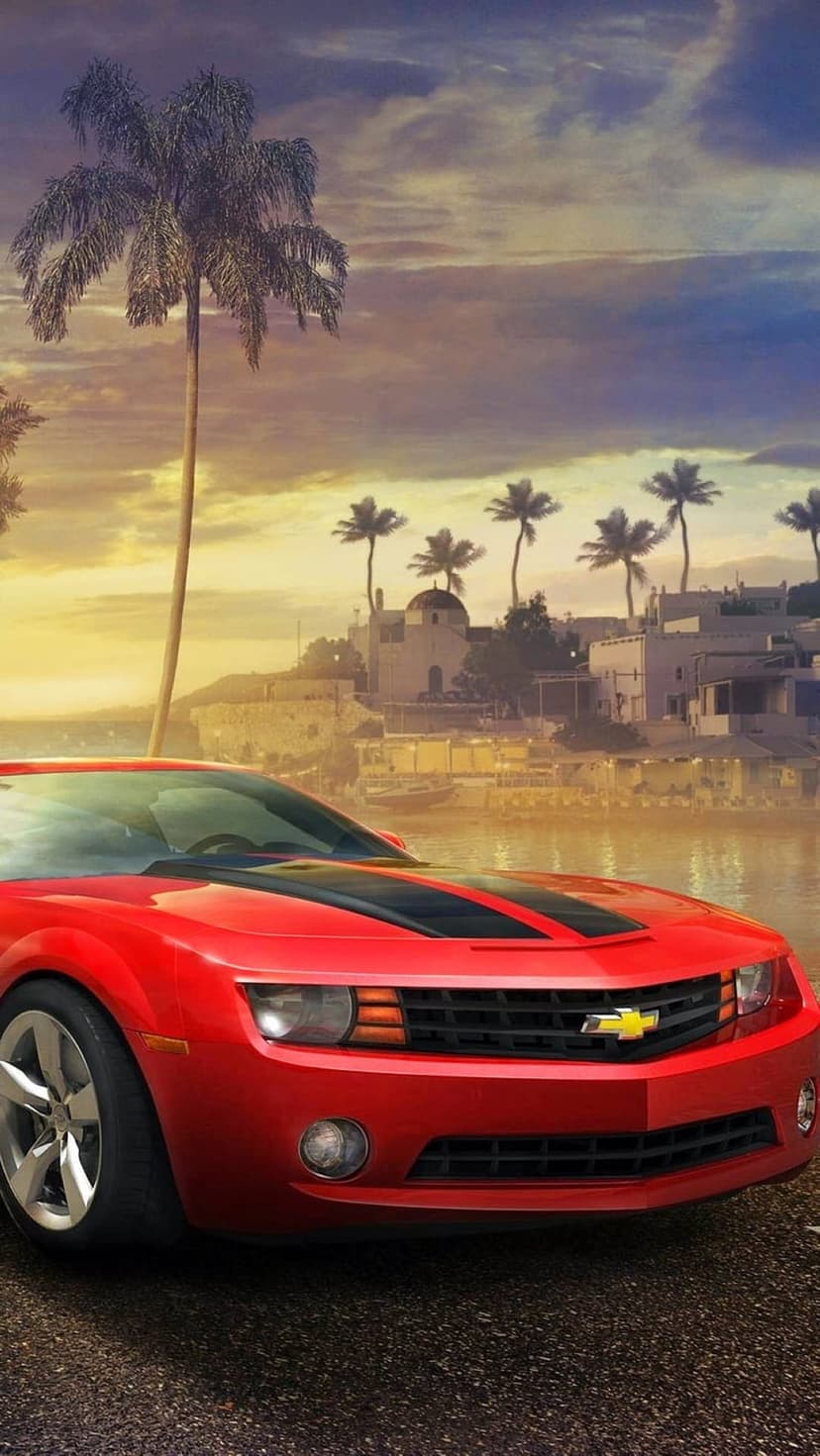 Red Chevrolet Camaro parked on a coastal road with palm trees and a village under a golden sunset sky.