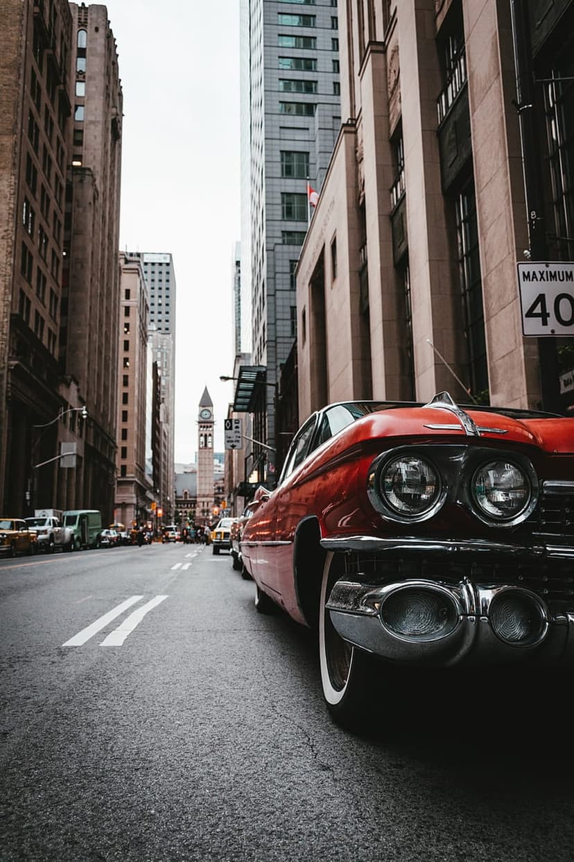 Red vintage Cadillac with chrome accents driving past tall skyscrapers on a busy metropolitan city street scene.