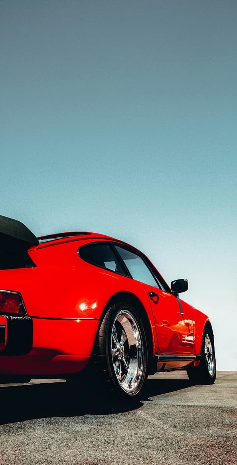 Red classic sports car with chrome wheels and a rear spoiler parked on a gray asphalt road under a blue sky.