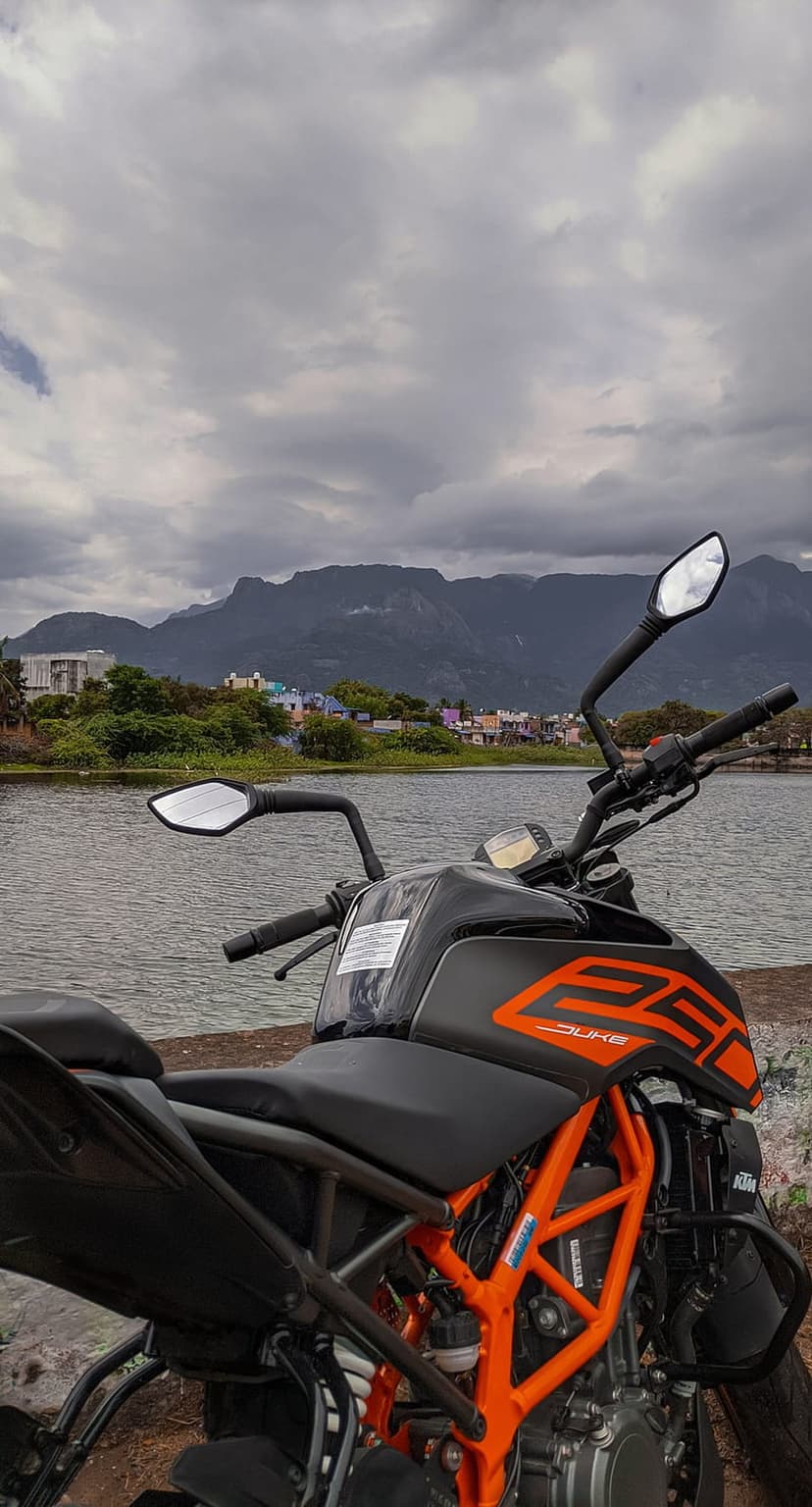 KTM Duke 250 motorcycle parked on a rocky lakefront with green mountains under a cloudy overcast sky.