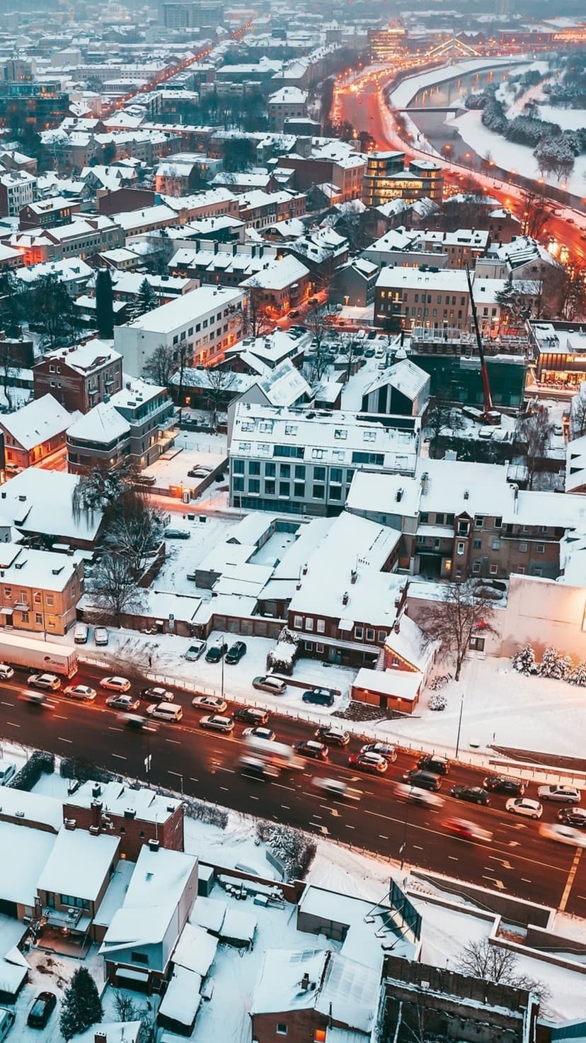 Snow-covered city rooftops and glowing streets with red light trails from traffic during a winter night.