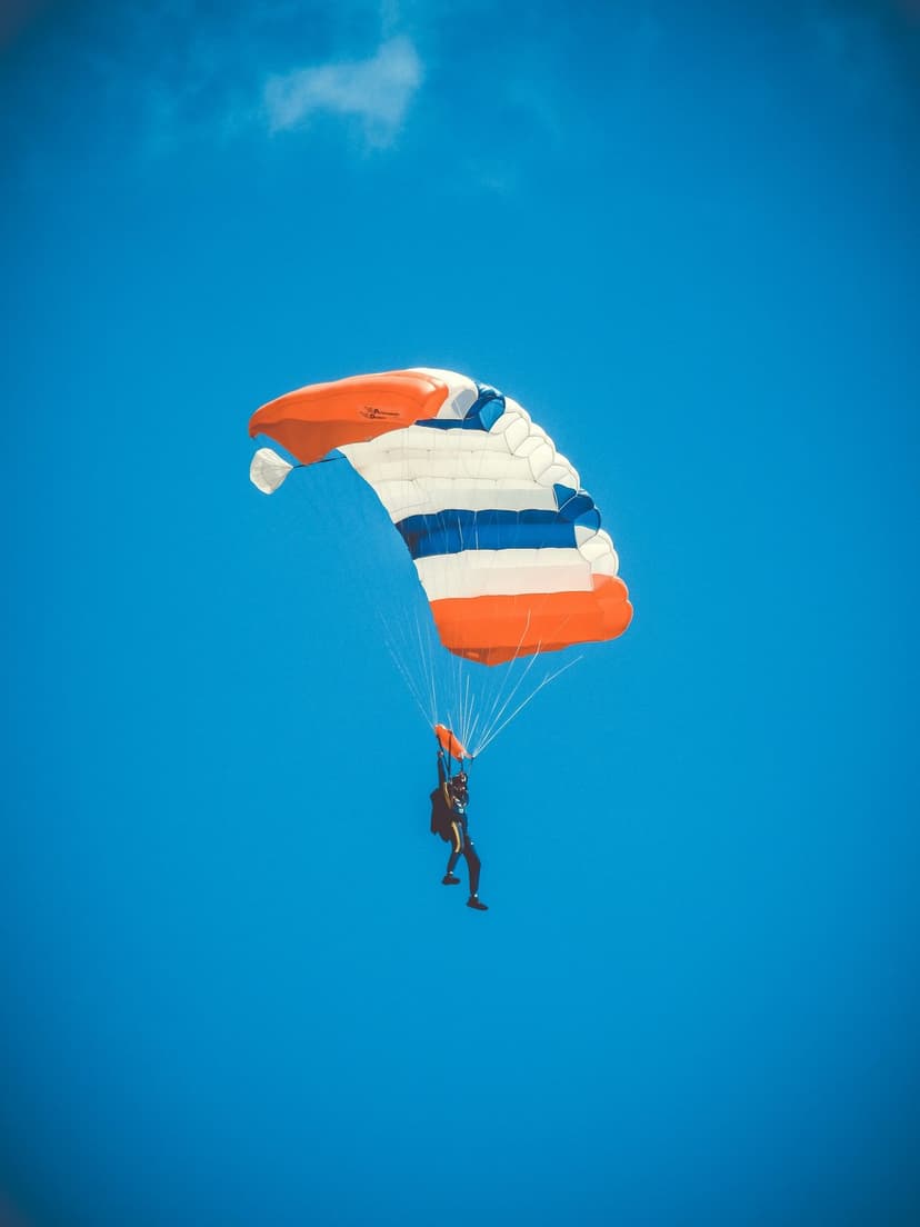 Skydiver descends through a clear blue sky beneath a large orange, white, and blue striped parachute.