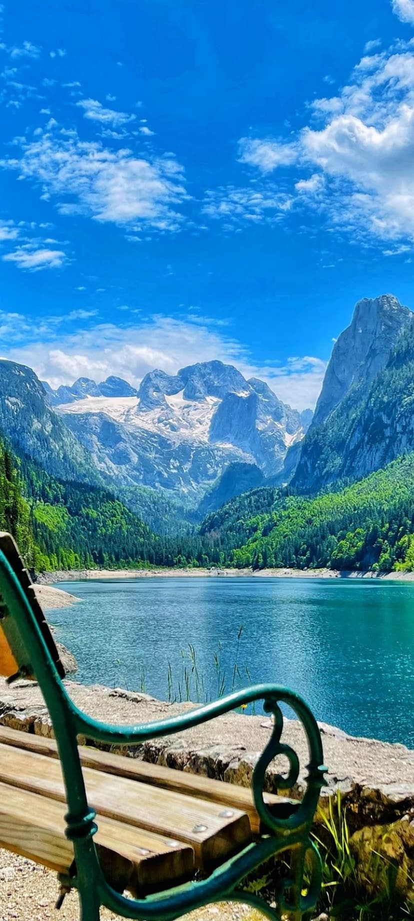 Weathered wooden bench faces a reflective mountain lake under snowy peaks and a bright blue sky with white clouds.