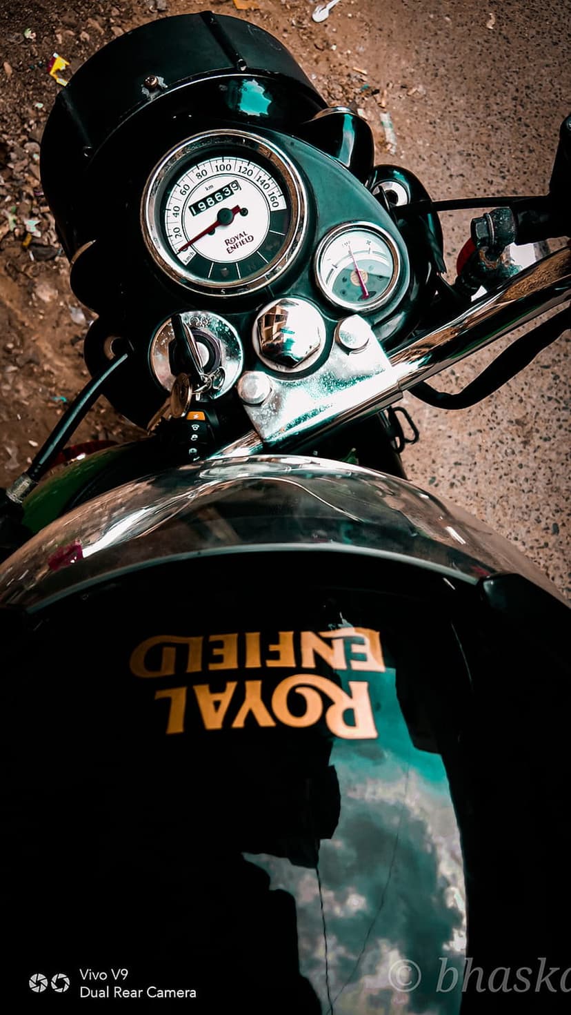Royal Enfield speedometer and fuel gauge featuring a gold logo on a black tank with soft sky reflections and chrome.