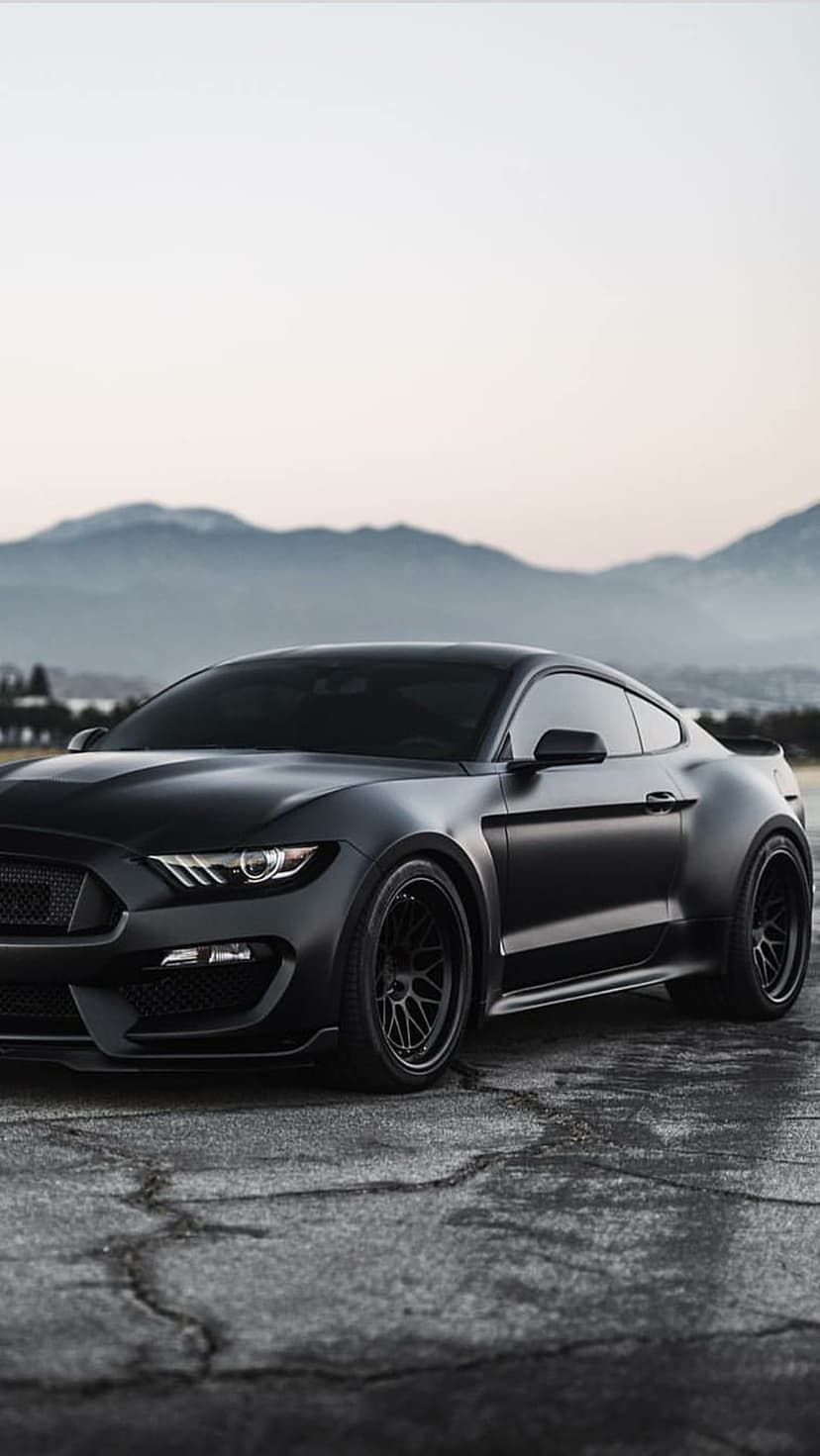 Matte black Ford Mustang GT on cracked asphalt with a scenic mountain range under a pale sky background.