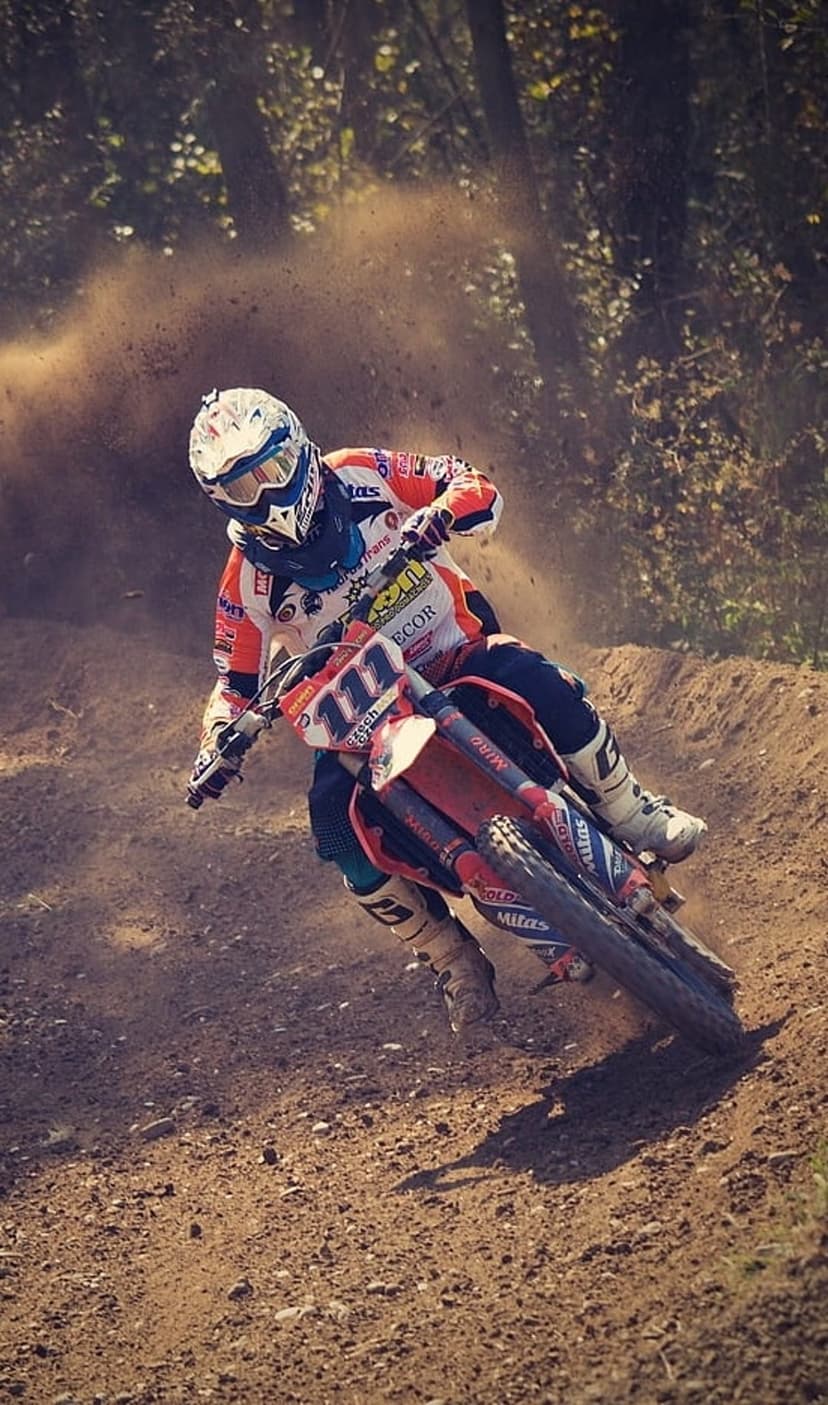 Motocross rider in orange gear leans a red dirt bike into a dusty turn on a sunlit racing track.