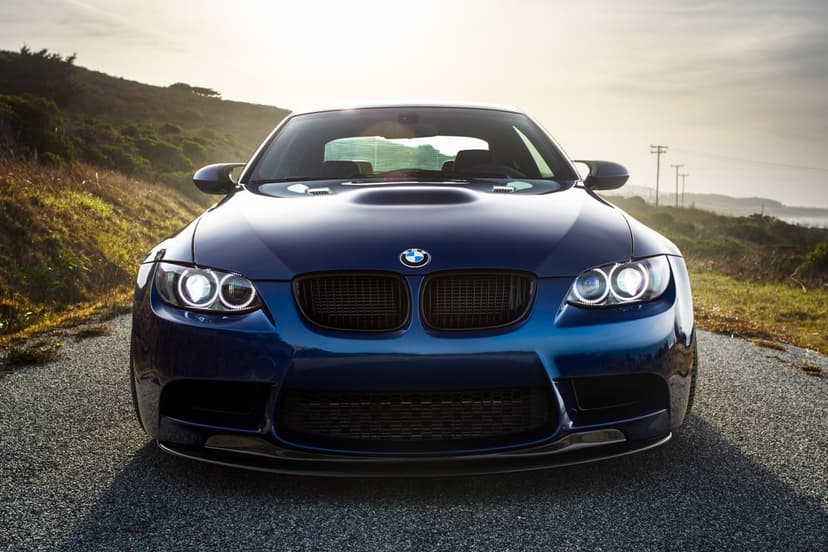 Blue BMW M3 sports car with halo headlights on a paved road with hills and sunset lighting in the background.