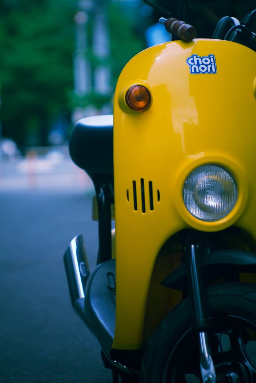Yellow scooter front end with a round chrome headlight and turn signal against a blurred green background.