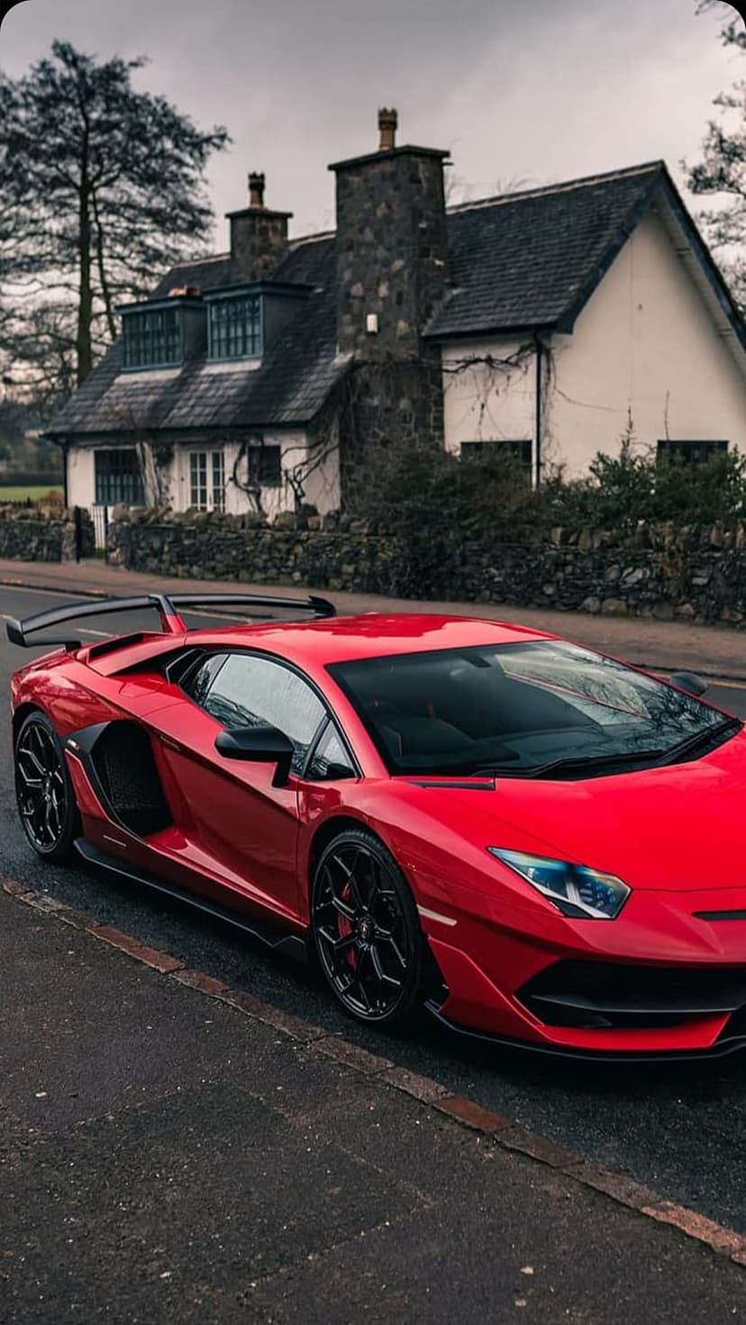 Red Lamborghini Aventador with a rear wing parked on a road beside a rustic white stone house and trees.