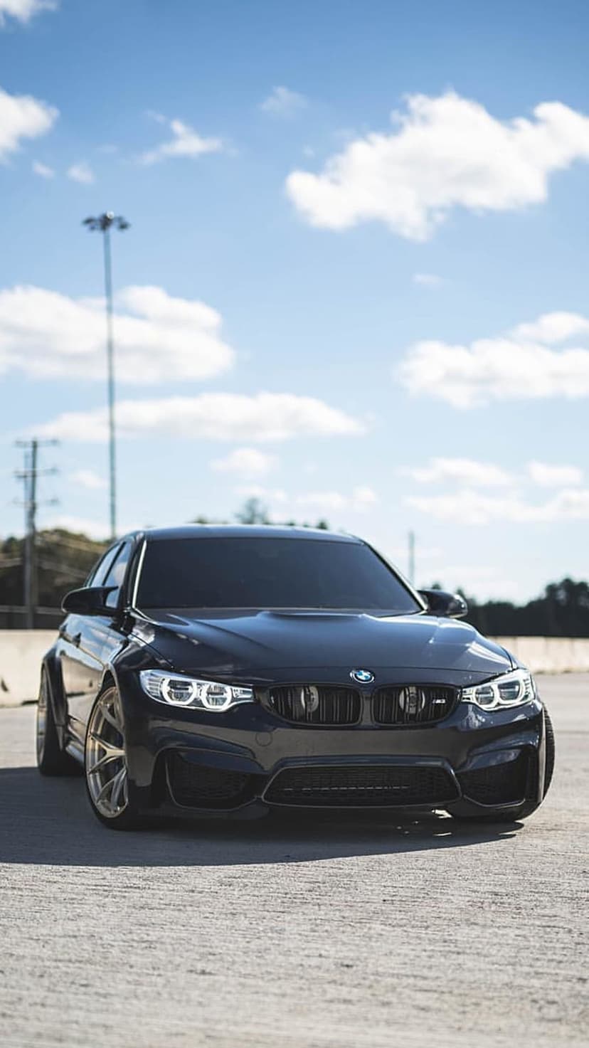 BMW M4 in shadow black paint parked on asphalt under a blue sky with clouds, highlighting front grille and rims.