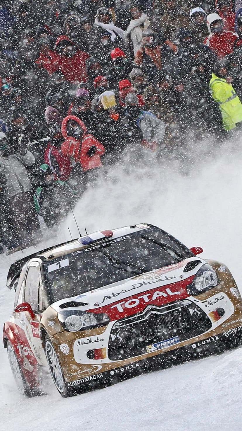 Citroen DS3 WRC rally car drifting through deep snow on a winter track with spectators and falling snowflakes.