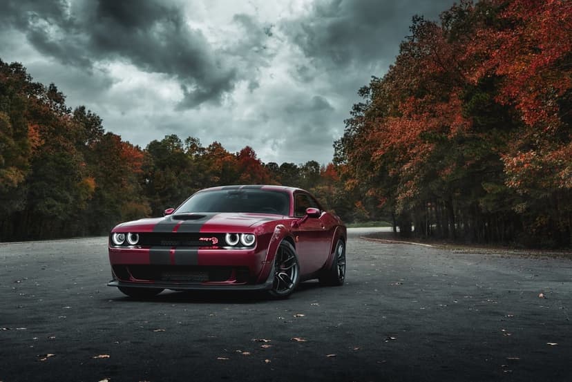 Red Dodge Challenger SRT Hellcat with racing stripes on a road surrounded by colorful autumn trees.